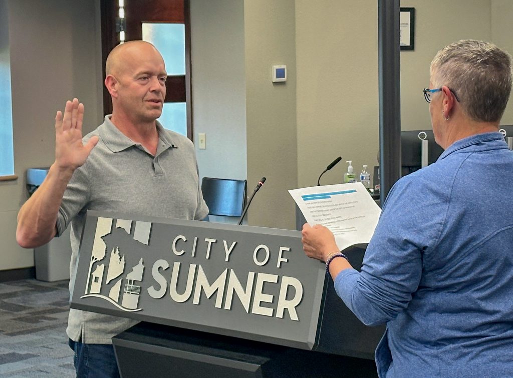 A man stands behind a City of Sumner podium with his right hand raised, being sworn in by a woman holding a document.