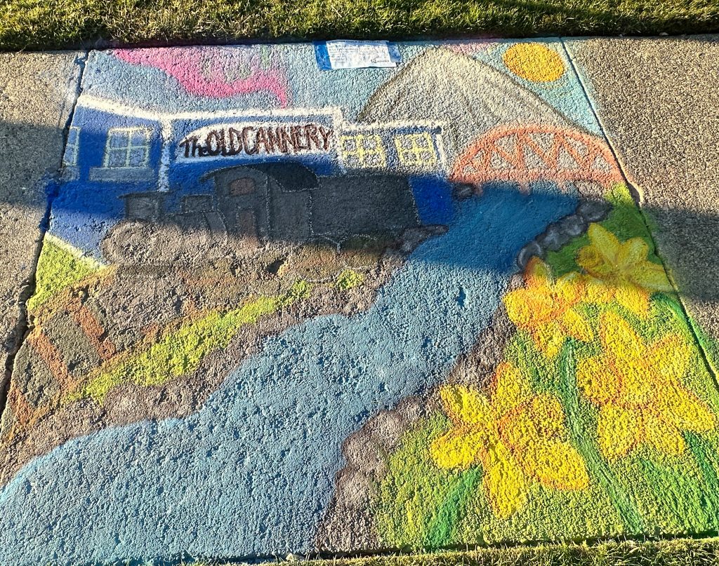 Chalk art on a sidewalk square depicting 'The Old Cannery' building, a mountain, a river, a red bridge, and large yellow daffodils.