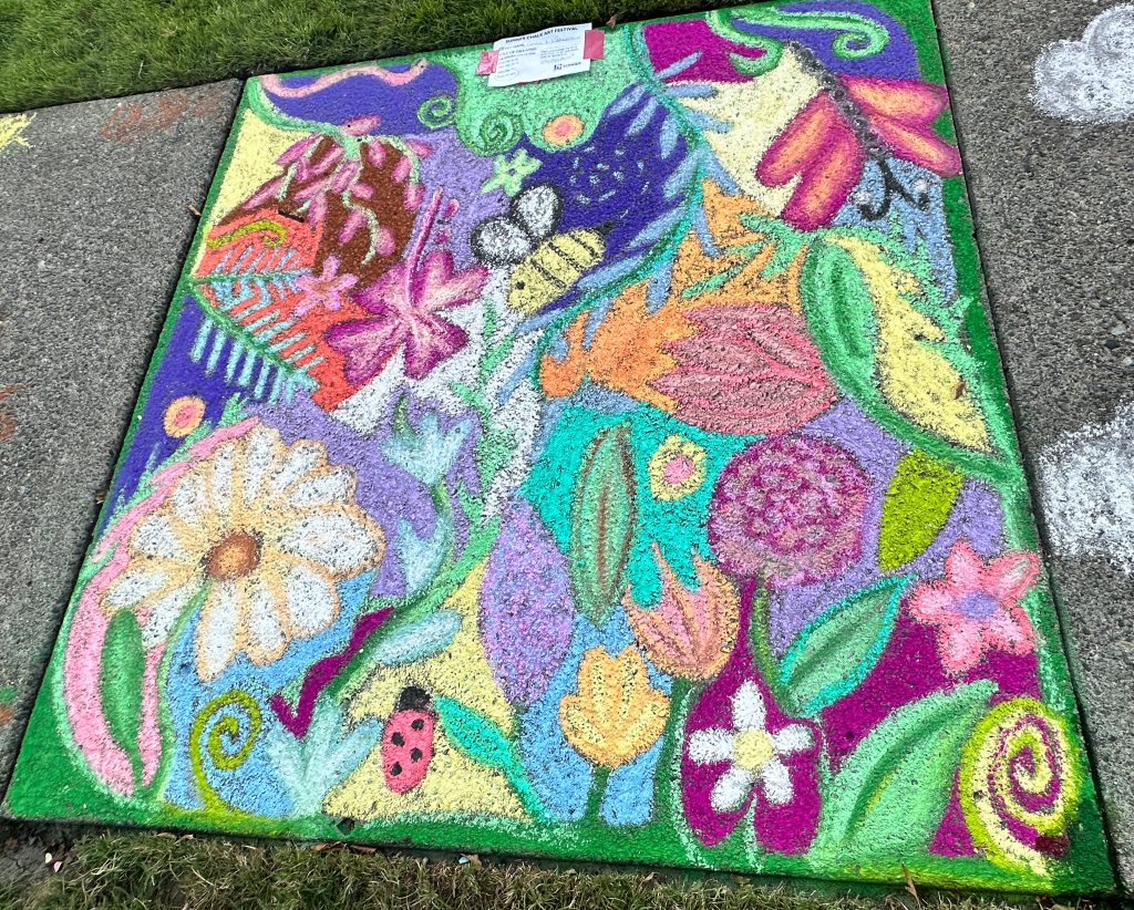 Colorful chalk art on a sidewalk square featuring a dense garden scene with flowers, a bee, a ladybug, a dragonfly, and vibrant foliage.
