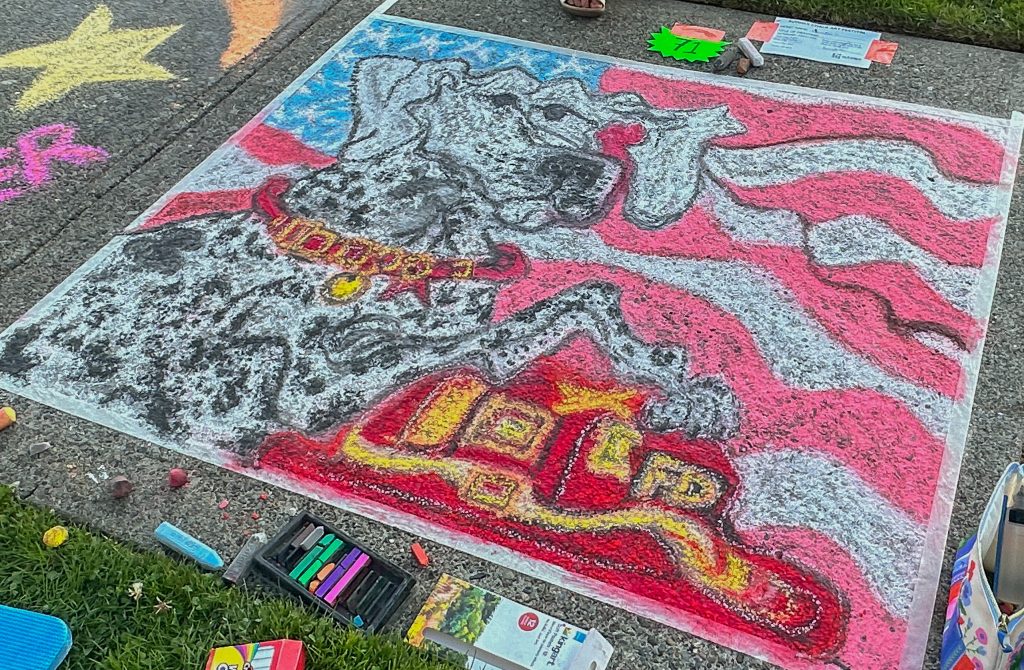 Sidewalk chalk art of a Dalmatian dog in front of a wavy American flag background, with a red fire truck at the bottom.